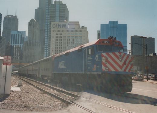 A blue Metra diesel locomotive numbered 115 pulls a line of silver passenger cars on a sunny day in downtown Chicago, with tall buildings and a street sign visible.