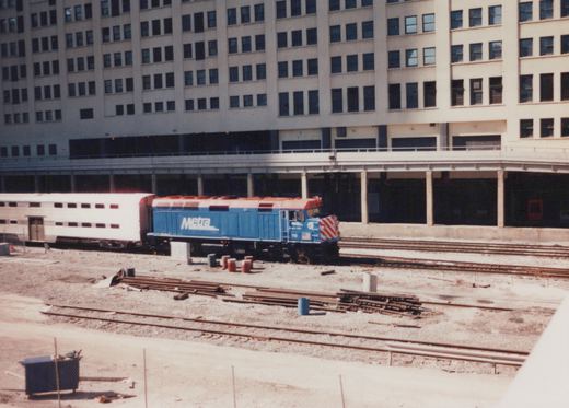A blue and red Metra locomotive numbered 112 is coupled to a white bi-level passenger car, stopped on tracks beside a tall building.