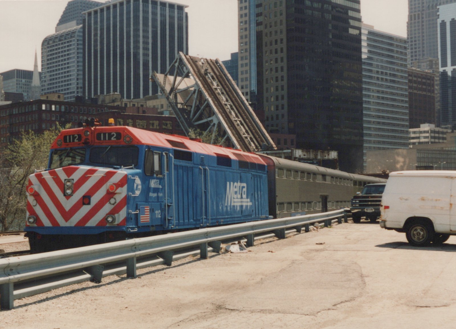 A blue and red Metra diesel locomotive numbered 112 pulls a line of stainless steel commuter cars on a track beside a road, with a raised drawbridge and Chicago skyscrapers in the background.