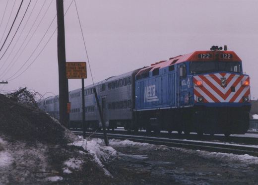 A blue and red Metra locomotive numbered 122 pulls a silver passenger train on a snowy day, seen from a three-quarter front view alongside a road with a yellow railroad sign.