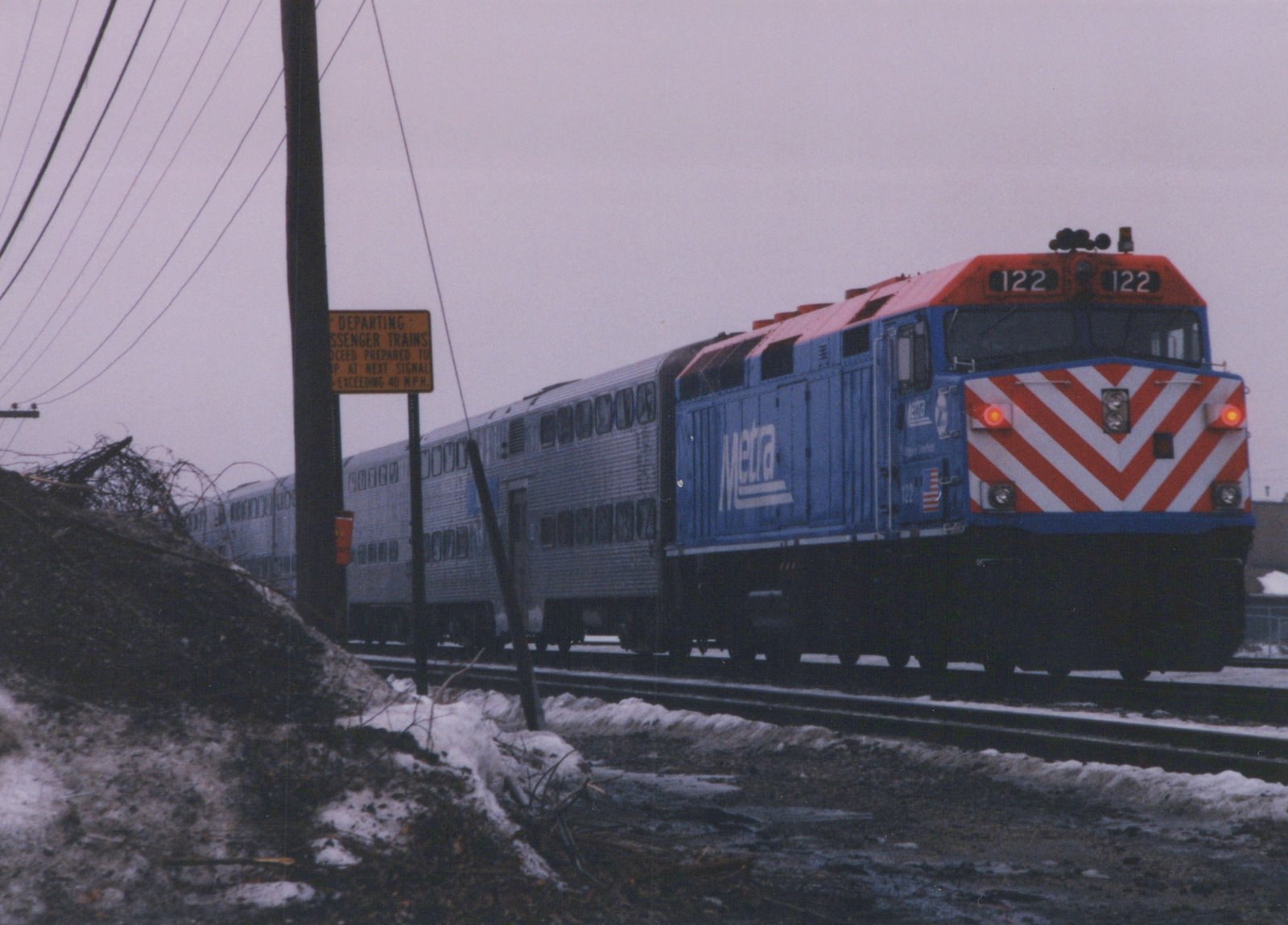 A blue and red Metra locomotive numbered 122 pulls a silver passenger train on a snowy day, seen from a three-quarter front view alongside a road with a yellow railroad sign.