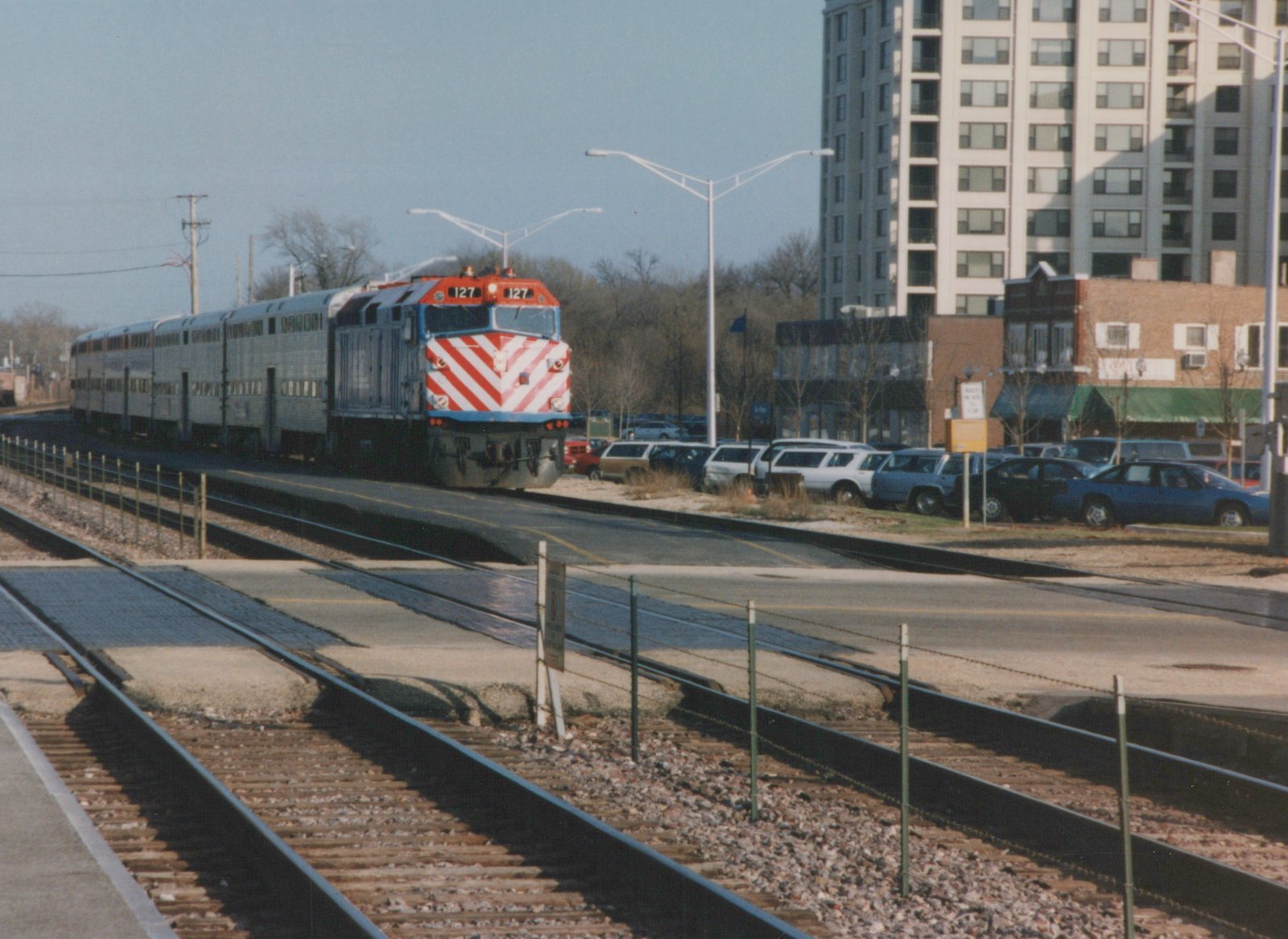 A red, white and blue striped Metra diesel locomotive numbered 127 leads a train of silver bi-level cars along tracks beside a road and a parking lot, with an apartment building in the background.