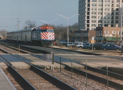 A red, white and blue striped Metra diesel locomotive numbered 127 leads a train of silver bi-level cars along tracks beside a road and a parking lot, with an apartment building in the background.