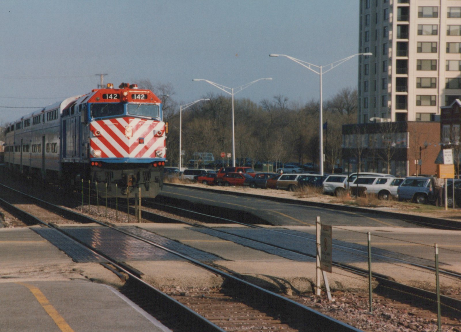A red, white, and blue Metra diesel locomotive numbered 142, pulling silver bi-level passenger cars, travels on tracks next to a street with parked cars and a tall building in the background on a clear winter day.
