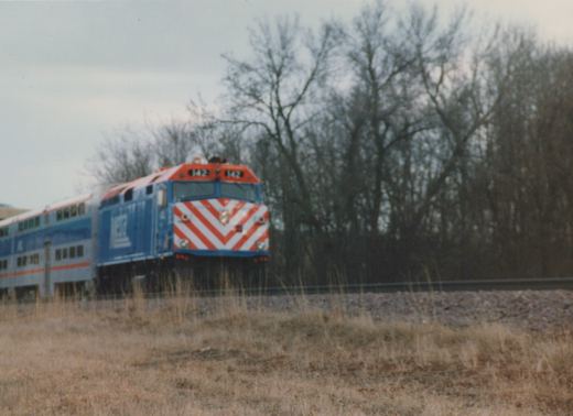A blue and red Metra locomotive numbered 142 pulls gallery cars along a track bordered by bare trees and dry grass under an overcast sky.