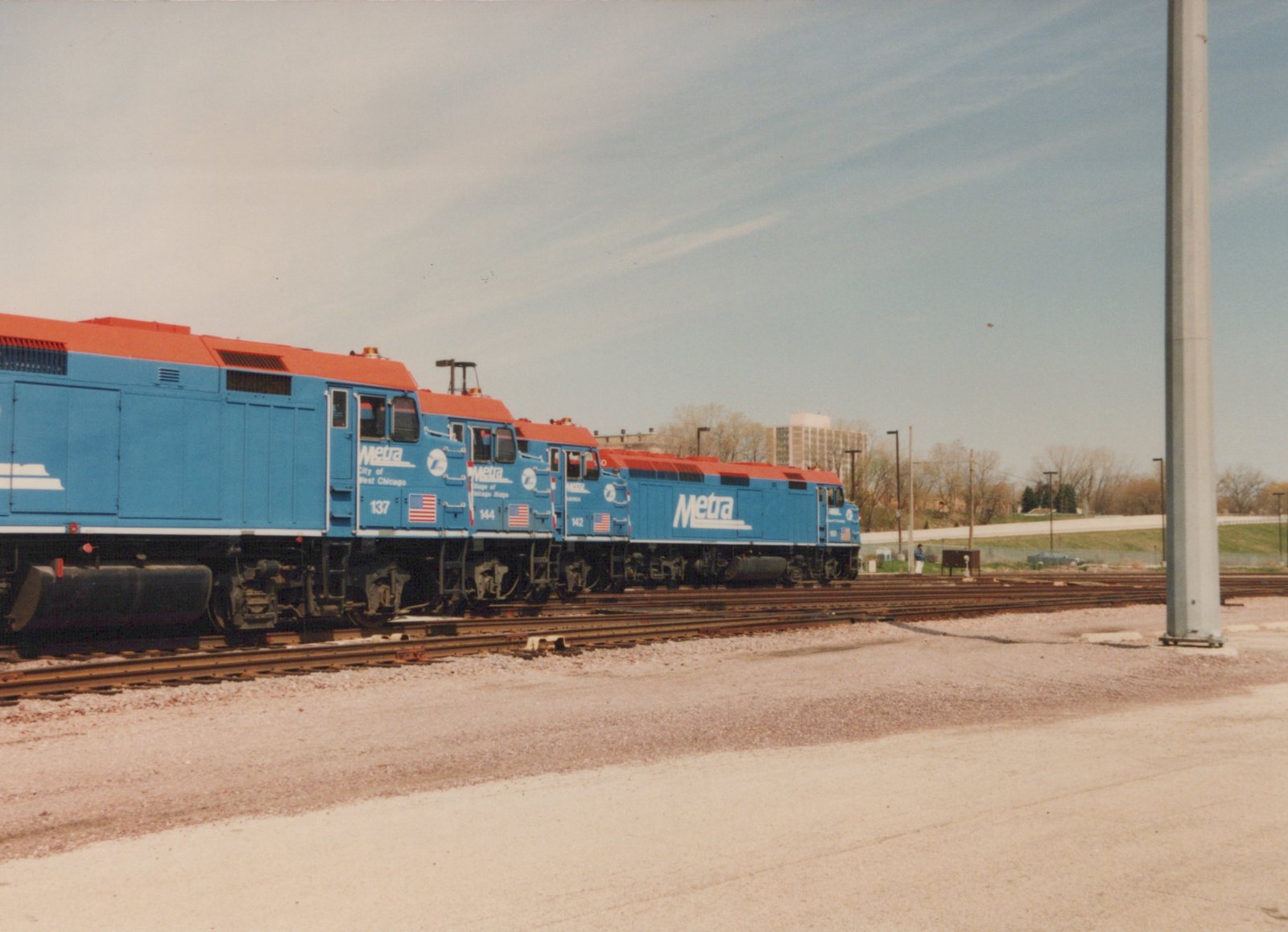 Three blue and red Metra locomotives with numbers 137, 144, and 142 are parked in a rail yard on a clear day, with a person standing nearby and buildings visible in the background.