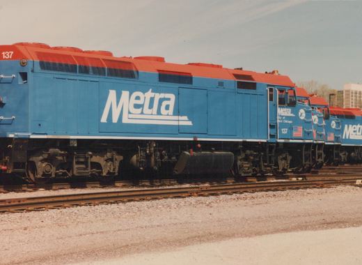 A blue and red Metra diesel locomotive numbered 137 is parked on parallel tracks in a rail yard under a clear sky.