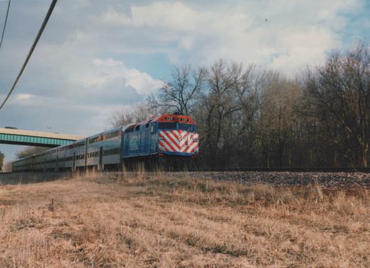 A blue, red and white Metra locomotive numbered 139 pulls a long train of silver double-decker cars along tracks beside a grassy embankment under an overcast sky, with leafless trees and a highway overpass in the background.