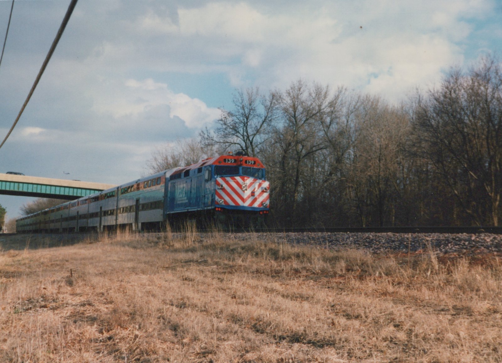 A blue, red and white Metra locomotive numbered 139 pulls a long train of silver double-decker cars along tracks beside a grassy embankment under an overcast sky, with leafless trees and a highway overpass in the background.