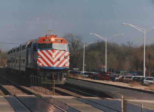A red, white, and blue Metra locomotive numbered 135 is moving on tracks beside a road with parked cars and bare trees.