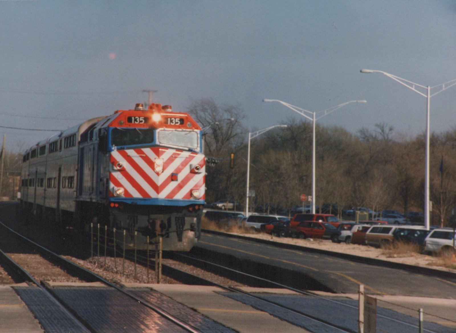 A red, white, and blue Metra locomotive numbered 135 is moving on tracks beside a road with parked cars and bare trees.