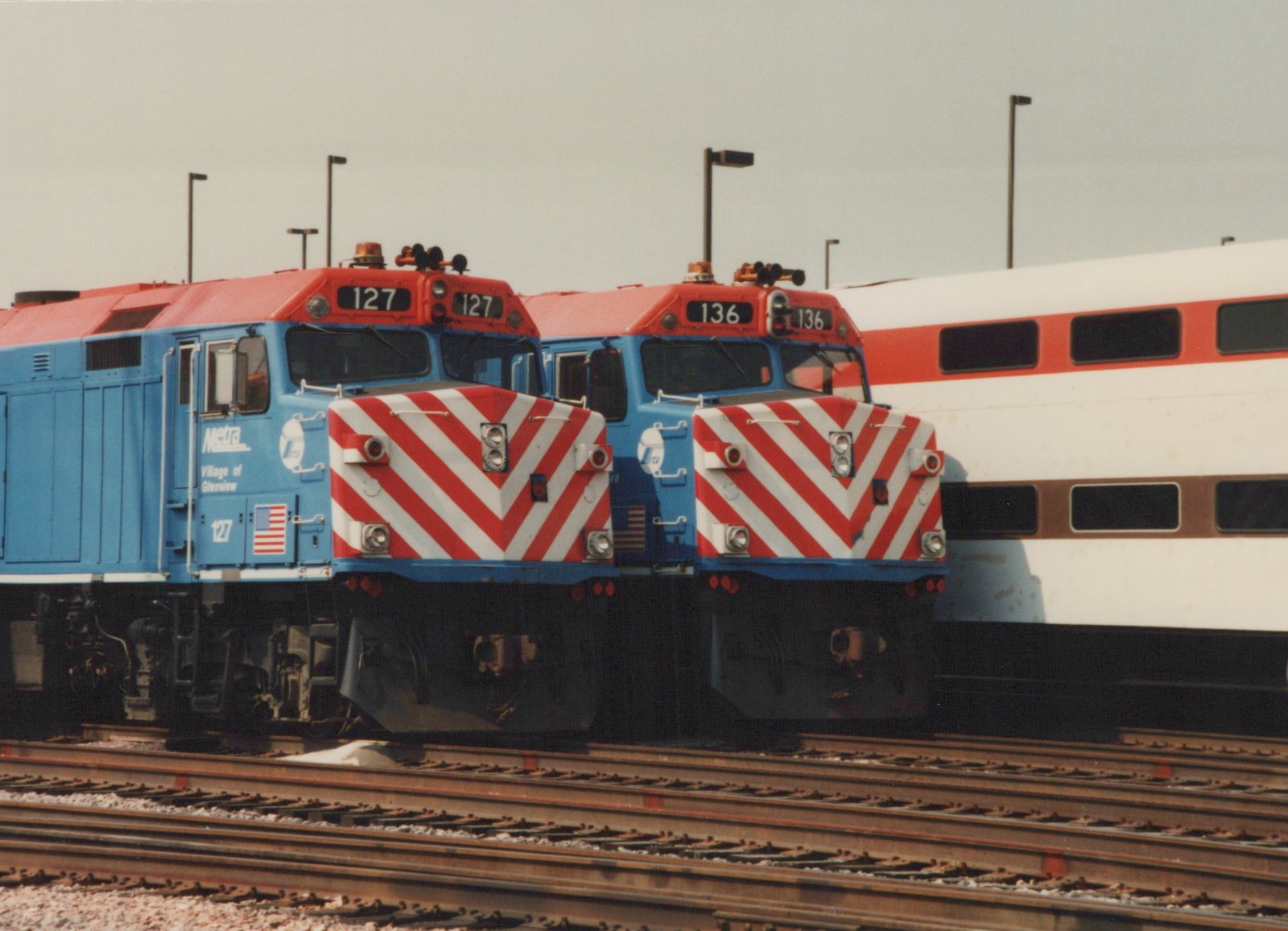 Two Metra diesel locomotives, 127 and 136, with red-and-white striped fronts, are parked on parallel tracks in a rail yard, with an Amtrak bi-level passenger car visible behind them.
