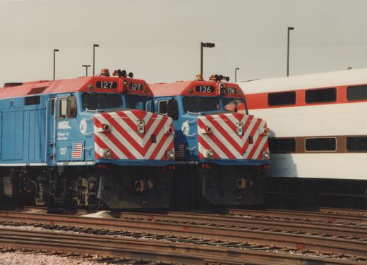 Two Metra diesel locomotives, 127 and 136, with red-and-white striped fronts, are parked on parallel tracks in a rail yard, with an Amtrak bi-level passenger car visible behind them.
