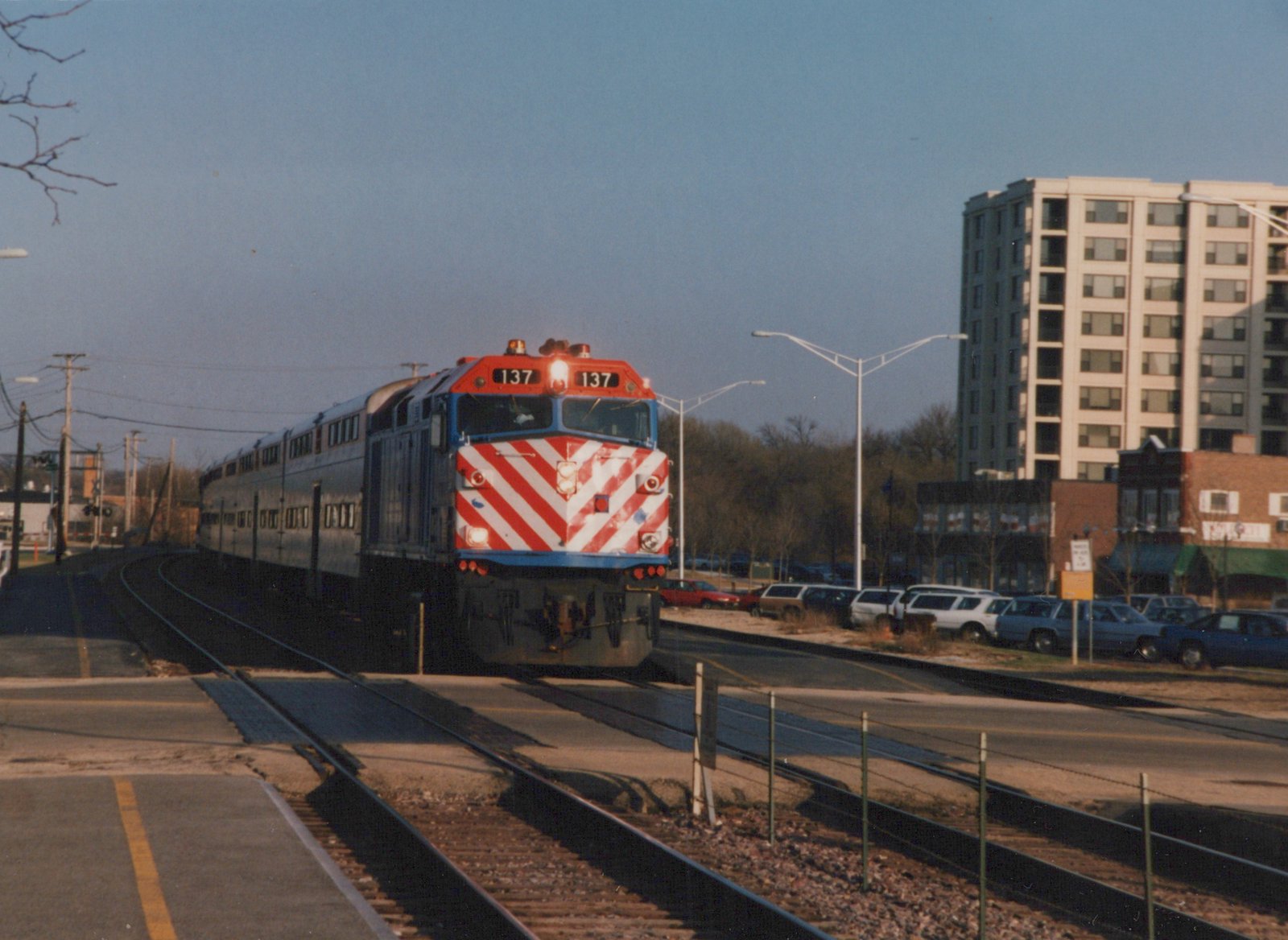 A red, white, and blue Metra locomotive with the number 137 on its front is moving along tracks in a suburban setting with buildings and parked cars nearby.