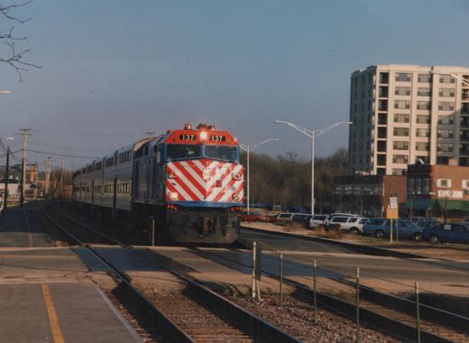 A red, white, and blue Metra locomotive with the number 137 on its front is moving along tracks in a suburban setting with buildings and parked cars nearby.