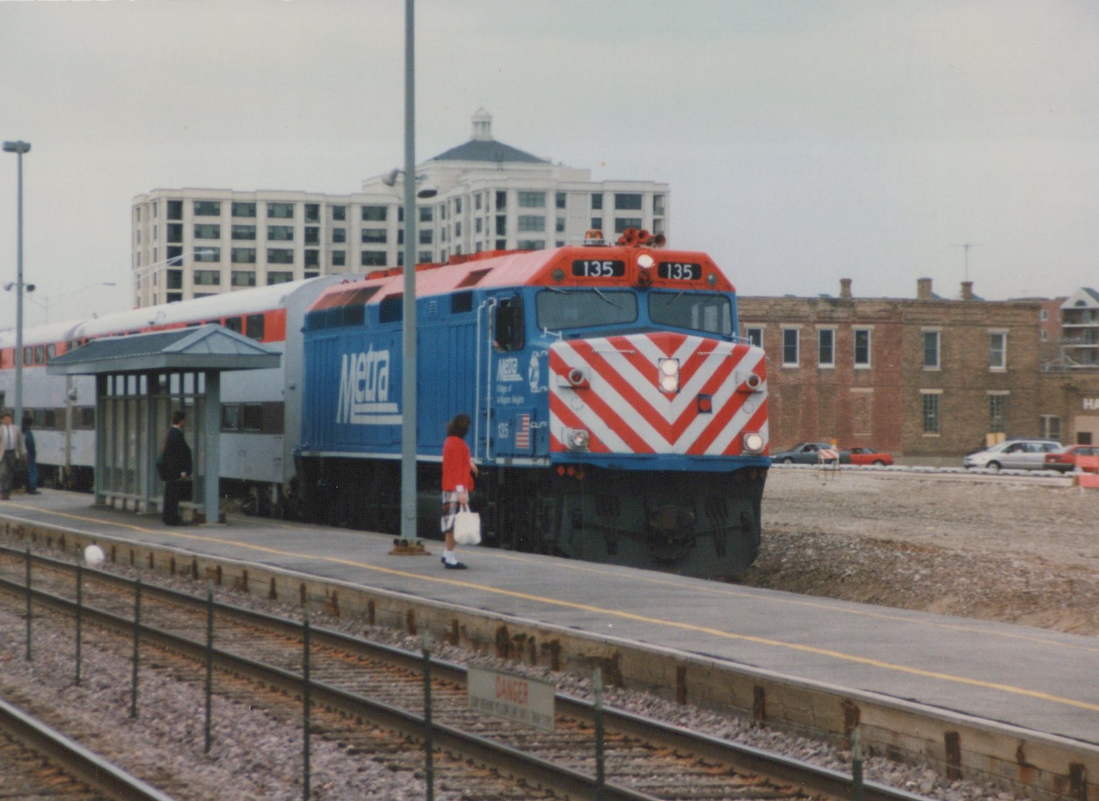 A blue and red Metra locomotive numbered 135 is stopped at a station platform. Several people are waiting, and a modern building is in the background.