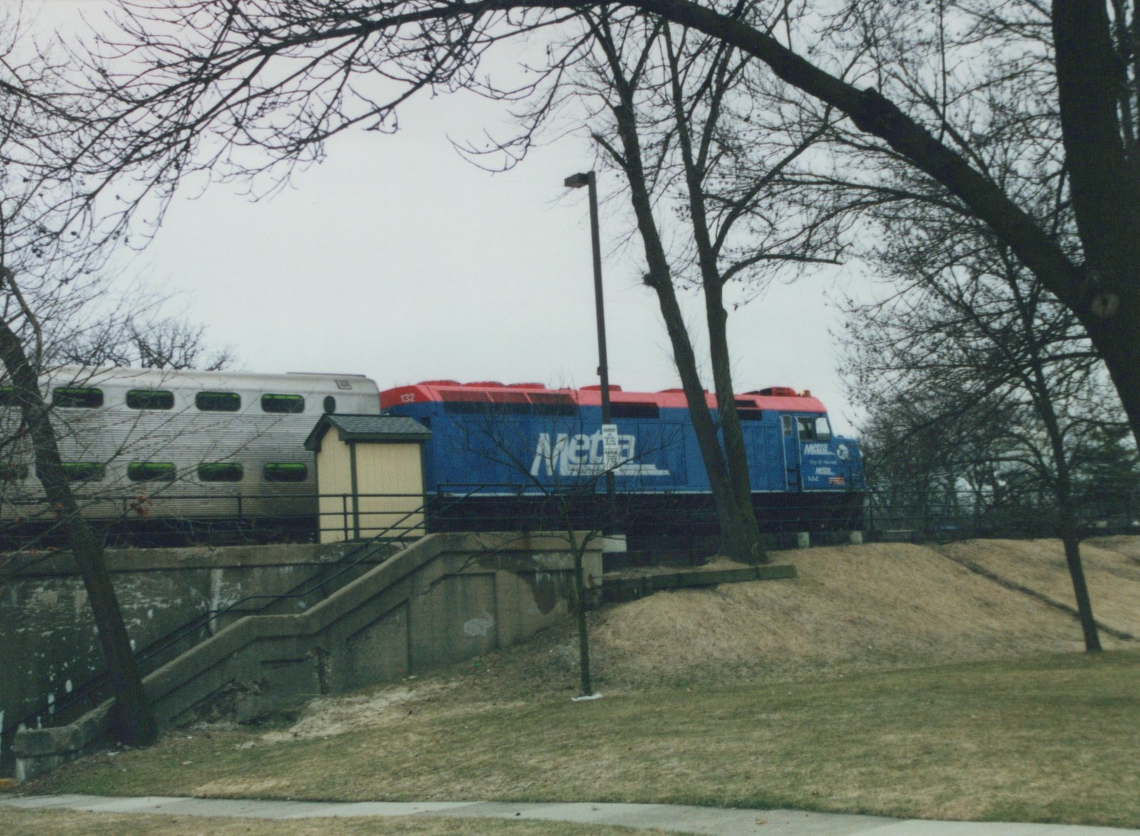 A blue and red Metra locomotive number 132 pulls a silver double-decker passenger car along an elevated track, viewed through bare winter trees.