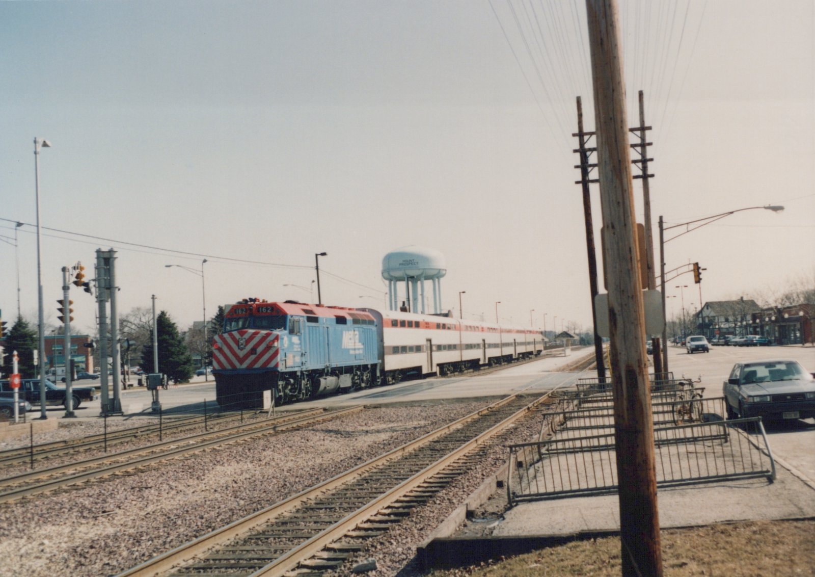 A blue and red Metra locomotive pulls a long train of white passenger cars across a street in a suburban area, with a water tower visible in the background.