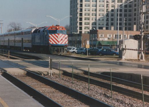 A blue, red, and white Metra locomotive numbered 160 pulls silver double-decker commuter cars along tracks next to a road with parked cars and a tall building in the background.