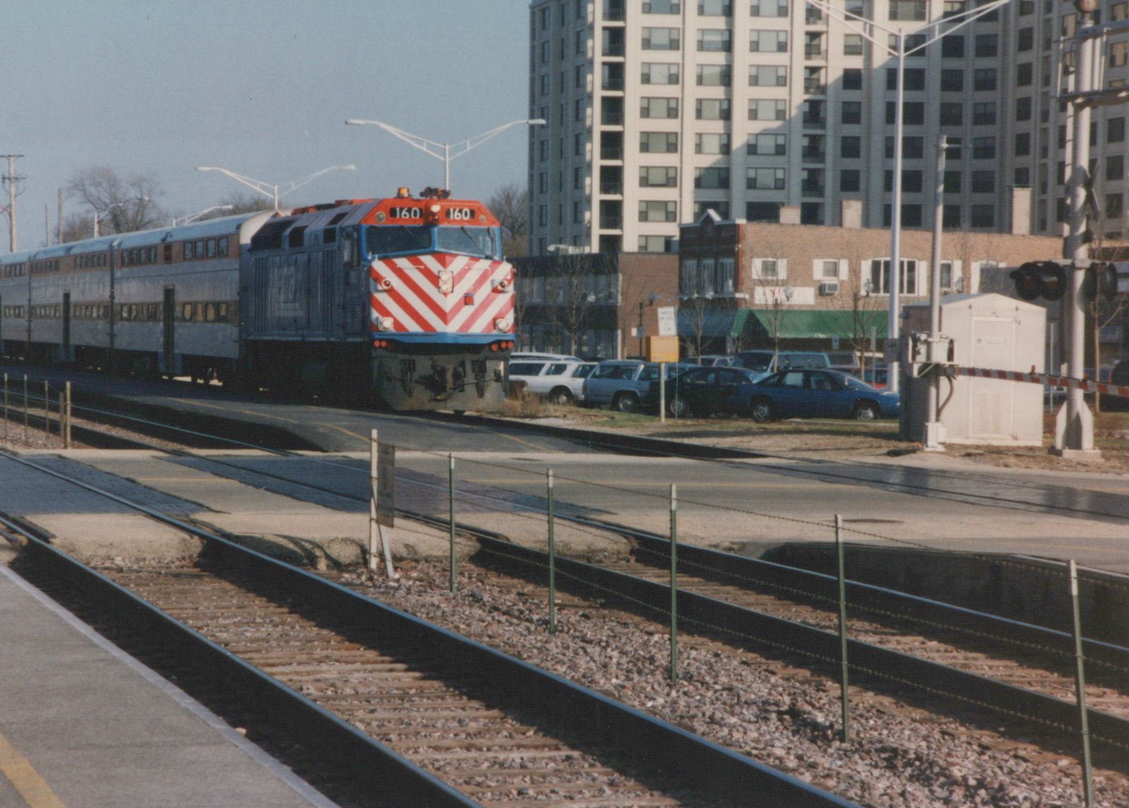 A blue, red, and white Metra locomotive numbered 160 pulls silver double-decker commuter cars along tracks next to a road with parked cars and a tall building in the background.