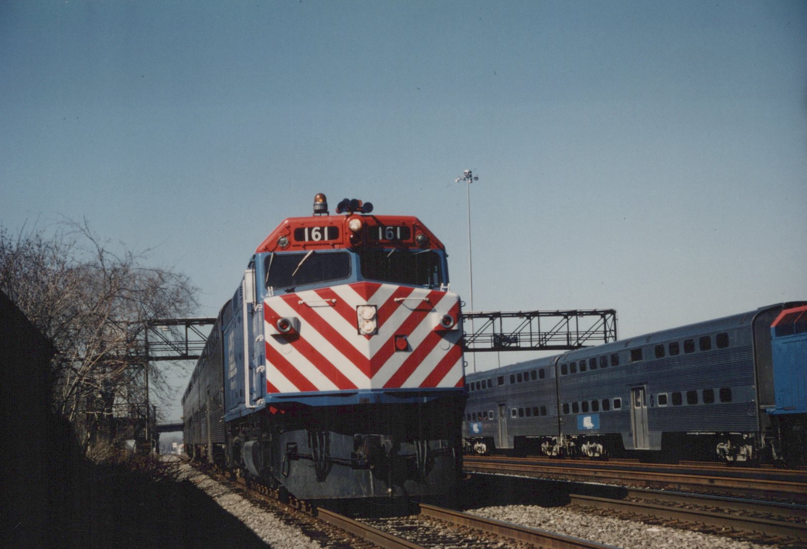A Metra train led by a blue locomotive with a red and white striped nose, number 161, pulling silver double-decker Gallery cars on a winter day with bare trees and clear sky.