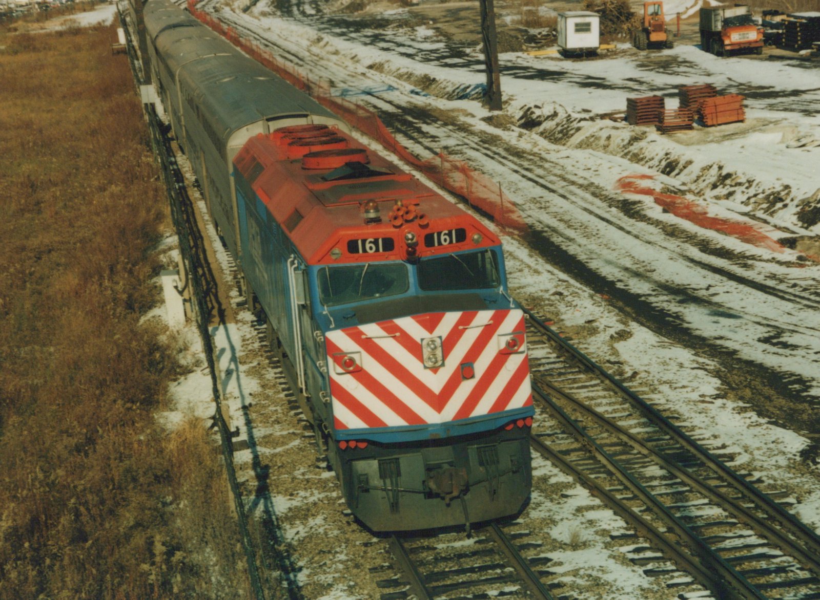 An overhead view of a blue and red Chicago and North Western locomotive pulling a line of double-decker commuter cars on tracks with snow on the ground.