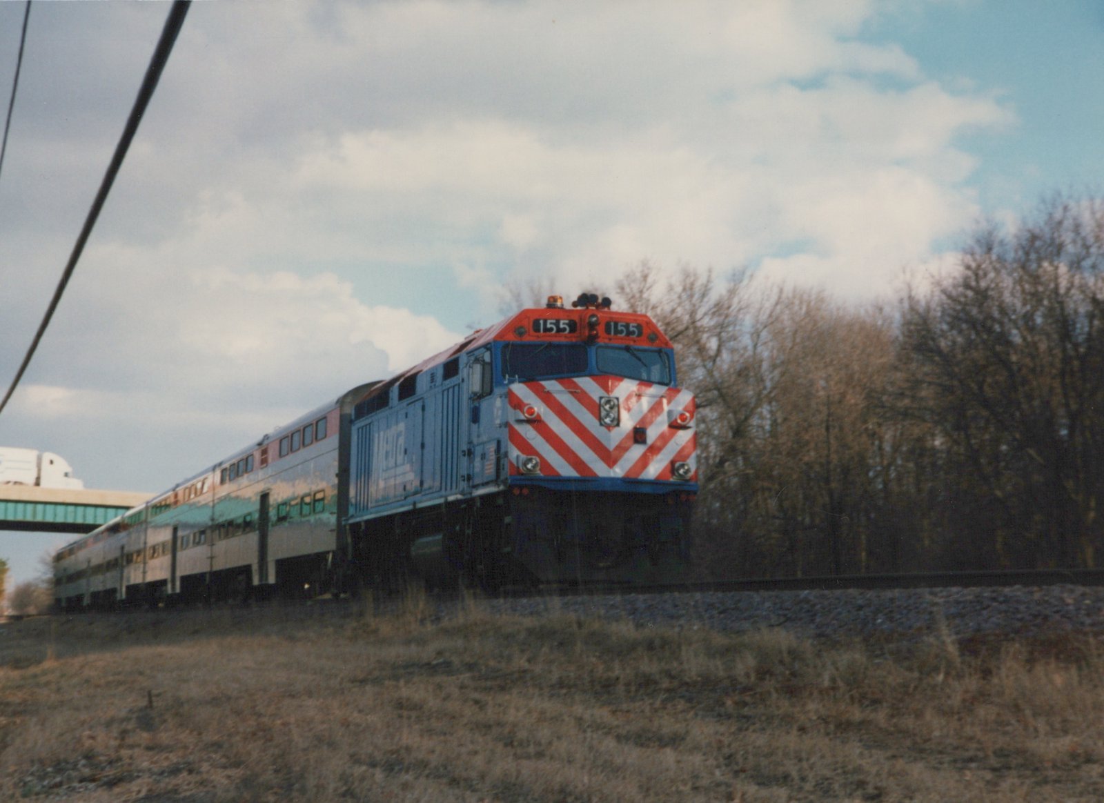 A blue and red Metra diesel locomotive pulls a train of double-decker cars along a tree-lined track on an overcast day.