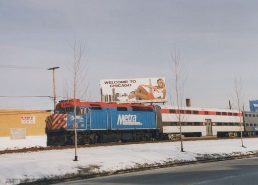 A blue and red Metra locomotive numbered 157, pulling white commuter cars, travels on a snowy track beneath a 'Welcome to Chicago' billboard.