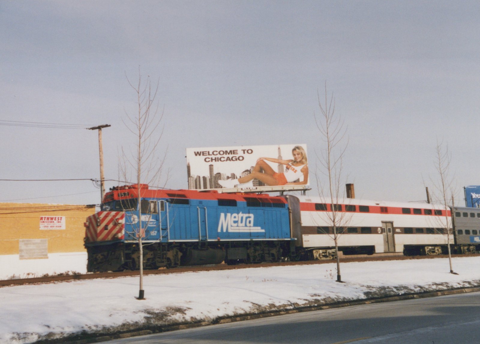 A blue and red Metra locomotive numbered 157, pulling white commuter cars, travels on a snowy track beneath a 'Welcome to Chicago' billboard.