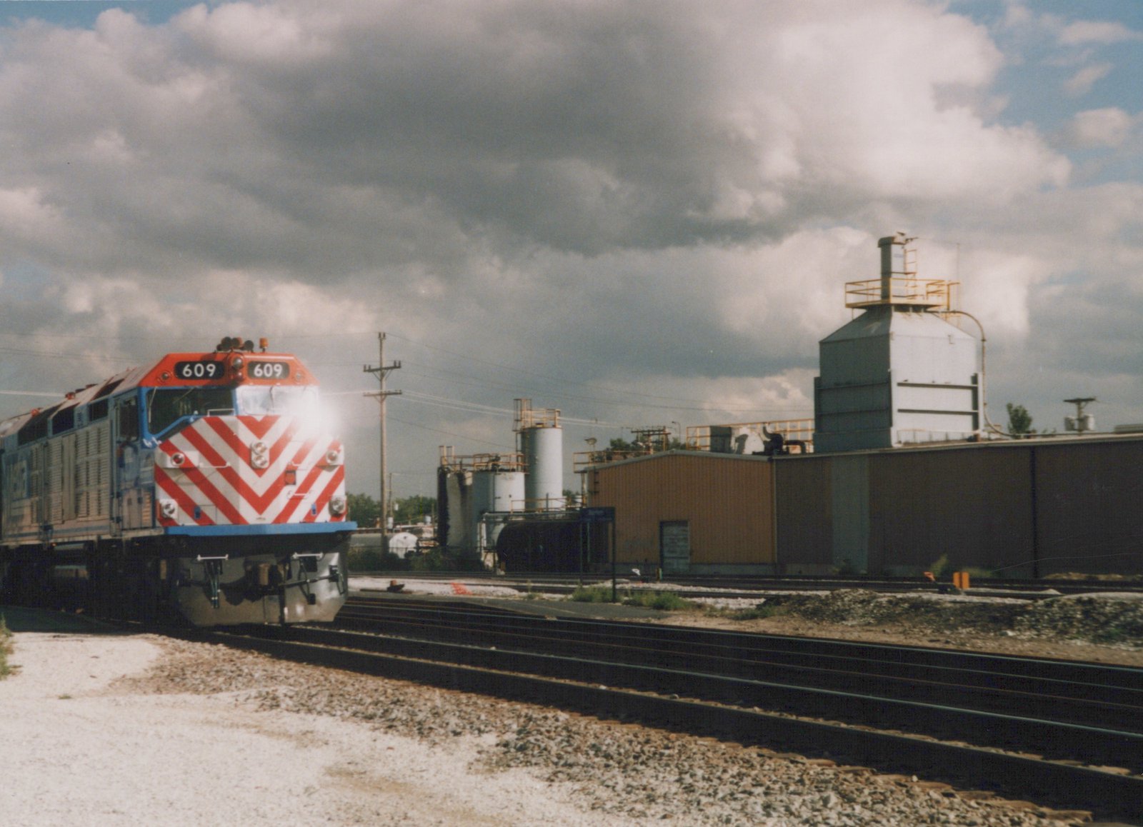A three-quarter front view of a Metra GP38-2 diesel locomotive numbered 609, painted in Metra's orange, white, and blue livery with a red and white chevron front, situated on a railroad yard with industrial buildings in the background under a cloudy sky.