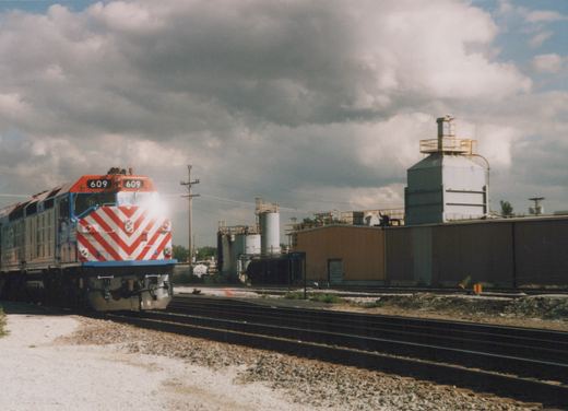 A three-quarter front view of a Metra GP38-2 diesel locomotive numbered 609, painted in Metra's orange, white, and blue livery with a red and white chevron front, situated on a railroad yard with industrial buildings in the background under a cloudy sky.
