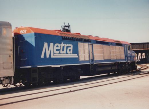 A blue and orange Metra locomotive numbered 607 is shown in profile, coupled to a silver passenger car, on railroad tracks near a bridge.