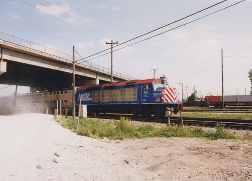 A blue and red Metra locomotive with the number 607 is pulling a line of stainless steel passenger cars under a large concrete bridge on a clear, sunny day.