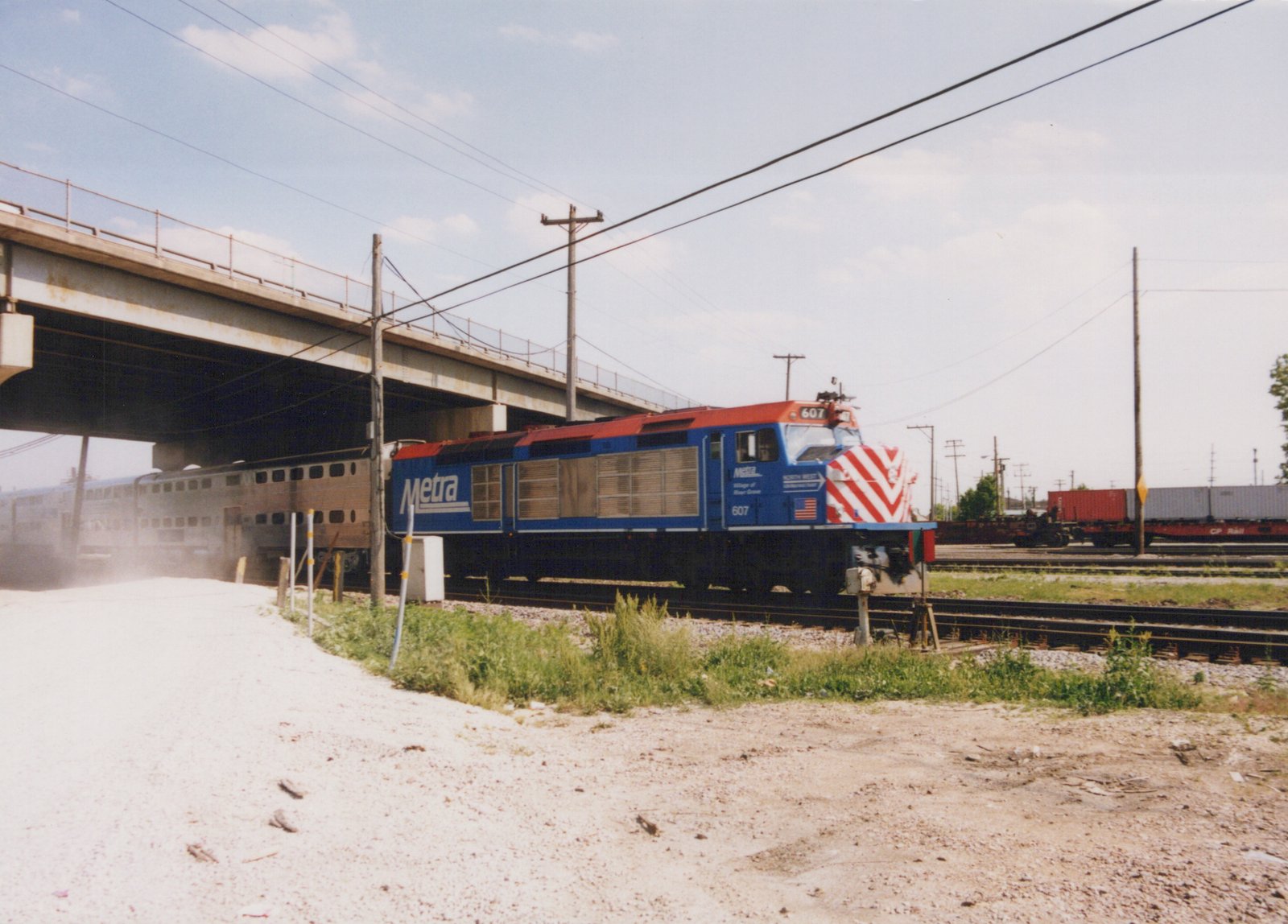 A blue and red Metra locomotive with the number 607 is pulling a line of stainless steel passenger cars under a large concrete bridge on a clear, sunny day.