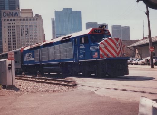 A blue Metra diesel locomotive, number 607, with a red and white striped nose, is on tracks in a city setting with tall buildings behind it.
