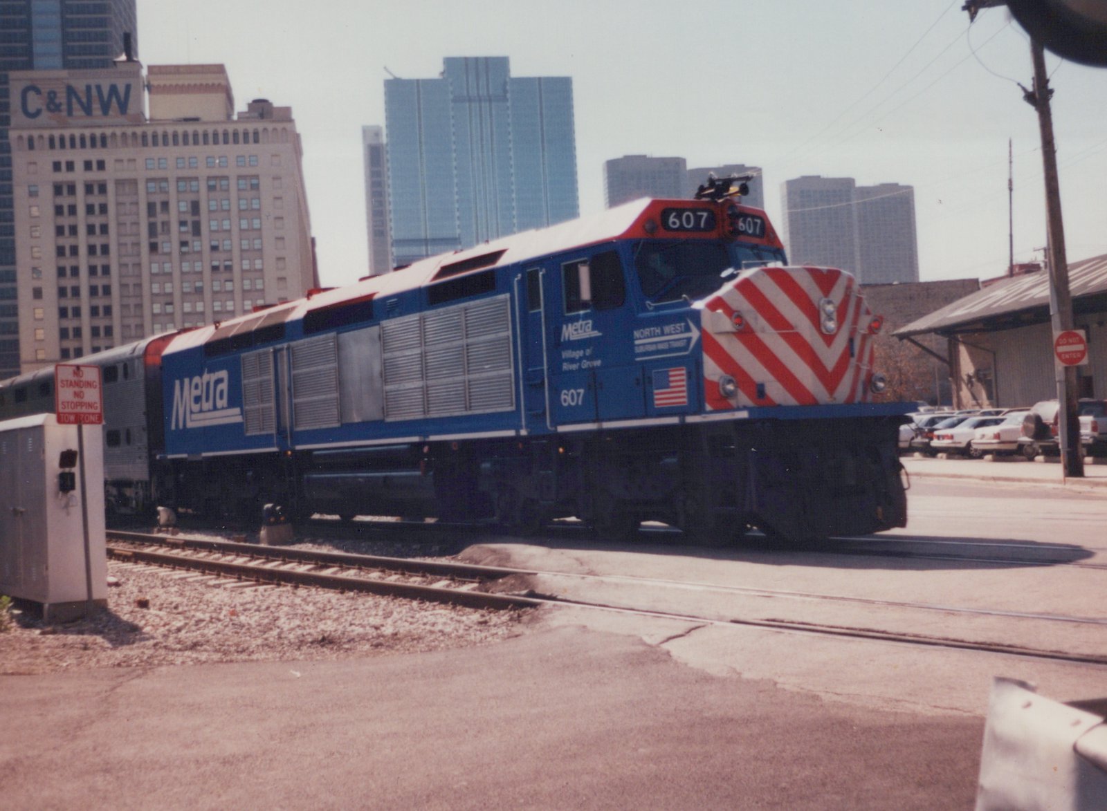 A blue Metra diesel locomotive, number 607, with a red and white striped nose, is on tracks in a city setting with tall buildings behind it.