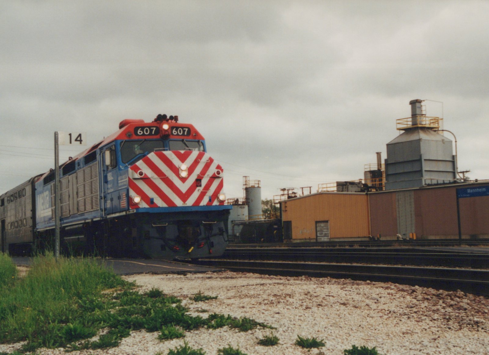 A blue, red, and white Metra locomotive numbered 607 is shown from a three-quarter front view, pulling a silver passenger car past a station named Mannheim. The sky is overcast, and industrial buildings are visible in the background.