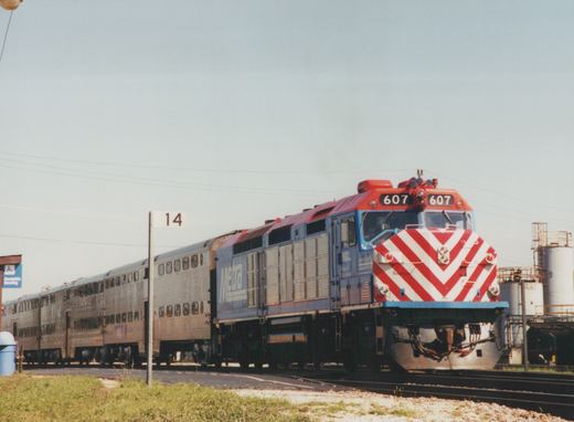 A blue and red Metra diesel locomotive with the number 607 pulls a line of silver double-decker passenger cars on a sunny day.
