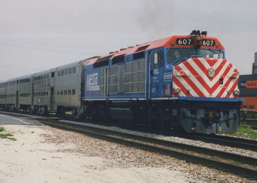 A blue and red Metra locomotive numbered 607 pulls a line of silver double-decker passenger cars on a railroad track under an overcast sky.