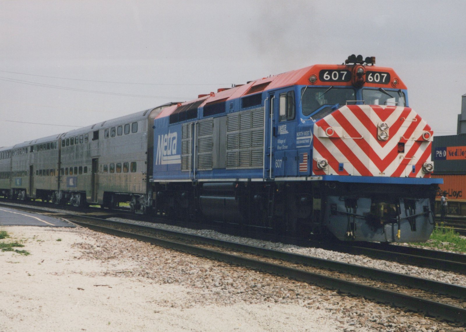 A blue and red Metra locomotive numbered 607 pulls a line of silver double-decker passenger cars on a railroad track under an overcast sky.
