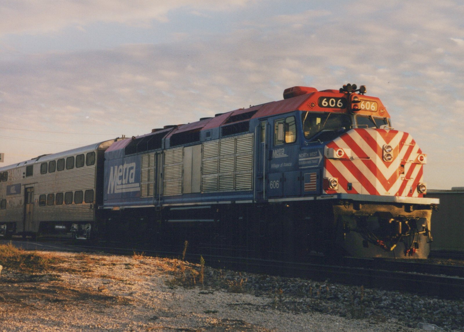 A blue Metra diesel locomotive with the number 606 and red and white chevron front, pulling a silver passenger car on a gravel track under a cloudy sky.