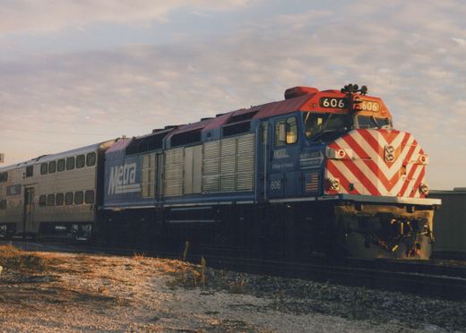 A blue Metra diesel locomotive with the number 606 and red and white chevron front, pulling a silver passenger car on a gravel track under a cloudy sky.