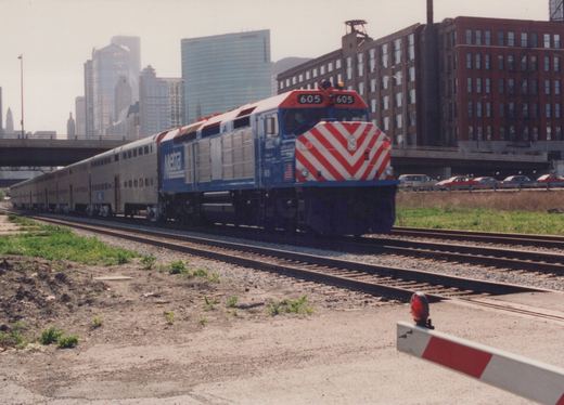 A blue, red, and white Metra diesel locomotive numbered 605 pulls a long train of silver passenger cars past a grade crossing, with a city skyline in the background.