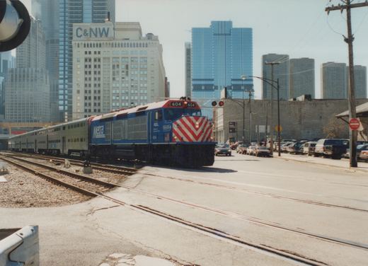 A blue Metra locomotive with the number 604 is pulling a train of silver commuter cars along tracks that cross a city street, with tall buildings in the background.