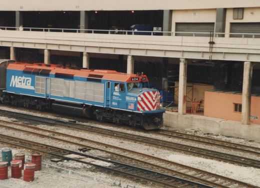 A blue and orange Metra diesel locomotive numbered 604 is parked on railroad tracks beneath an elevated concrete structure, with red and white striped front buffers and a person visible in the cab.
