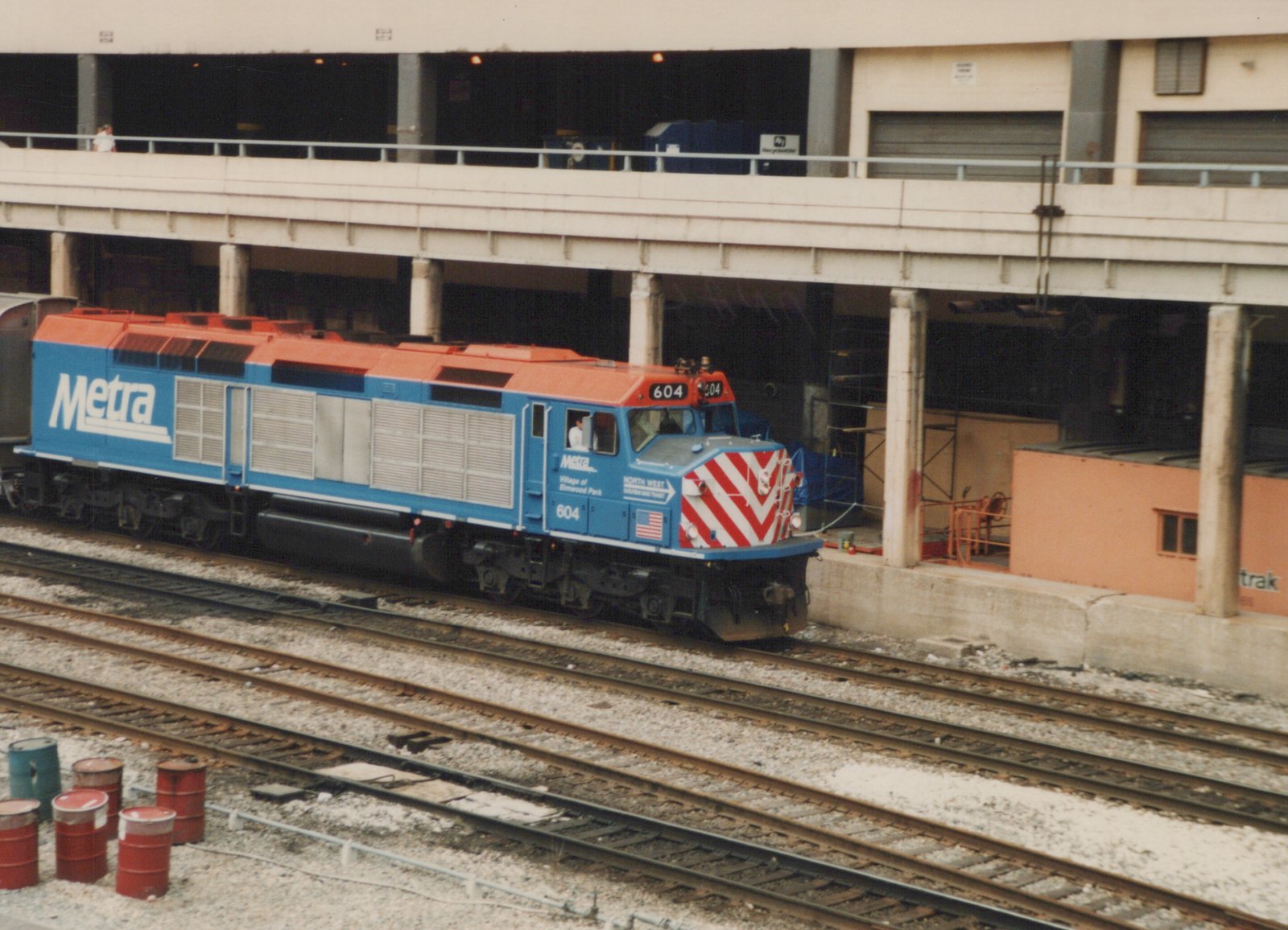 A blue and orange Metra diesel locomotive numbered 604 is parked on railroad tracks beneath an elevated concrete structure, with red and white striped front buffers and a person visible in the cab.