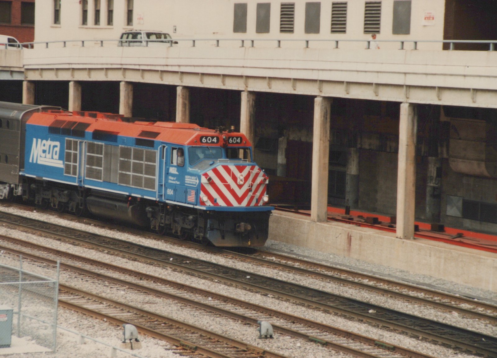 A blue and orange Metra locomotive, number 604, pulling a double-decker passenger car under an elevated highway in Chicago.