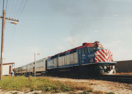 A blue and red Metra locomotive numbered 603 leads a silver train of double-decker gallery cars on a railroad track beside a grassy area, with a person standing near a small shelter in the background.