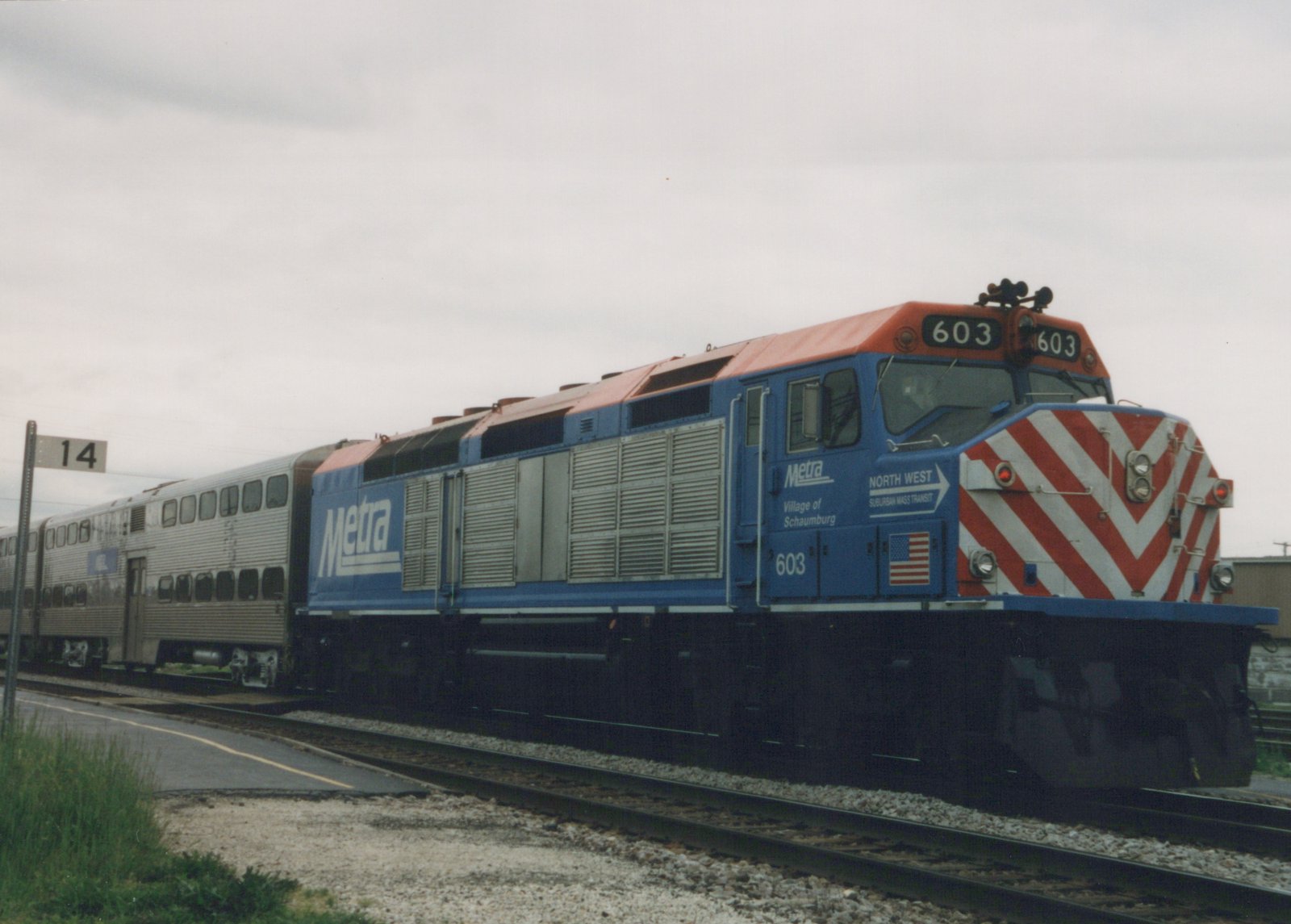 A blue Metra locomotive with the number 603 on the front and sides, pulling a train of silver bi-level passenger cars on a railroad track under an overcast sky.