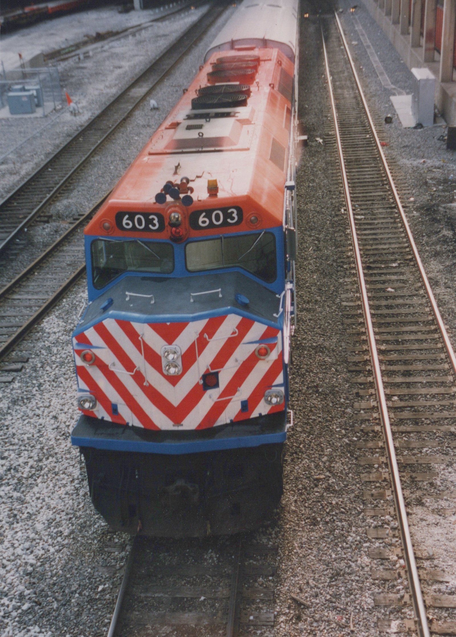 A front view of a Metra EMD F40PH locomotive, number 603, sitting on tracks in a yard, painted in orange, blue, and gray with red and white chevron stripes on the front.
